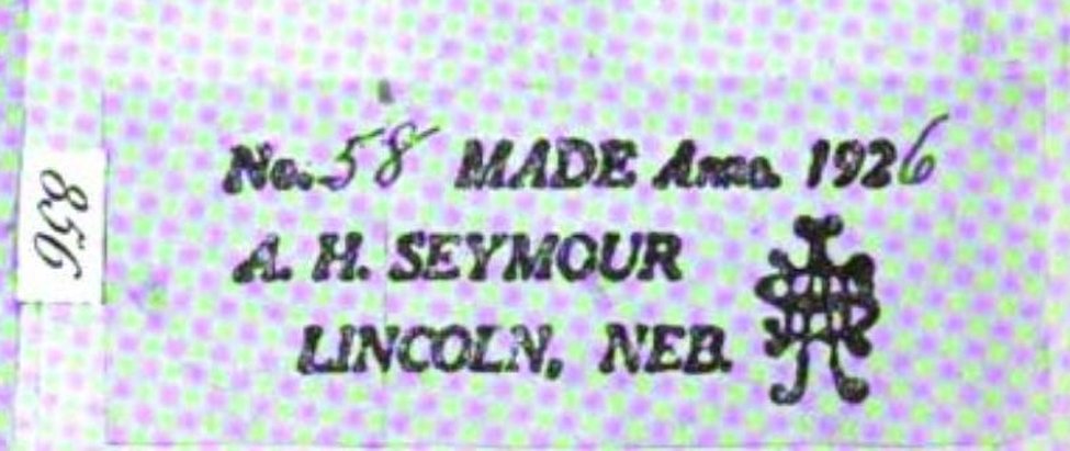 A printed violin label in a bold slanted face, shown turned a quarter-turn clockwise from the plate, where it is printed on its side. Three lines read No. 58 MADE Anno 1926; then A. H. SEYMOUR; then LINCOLN, NEB. The serial 58 and the final digit of the year are heavier and less regular than the type around them, having been filled in by hand. At the right stands a small dense monogram device of interlaced letters. At the left edge the plate's facsimile number, 856, sits in its own small ruled box and remains on its side, because only the label has been turned. A coarse halftone dot screen from the 1930 printing lies over the whole image.