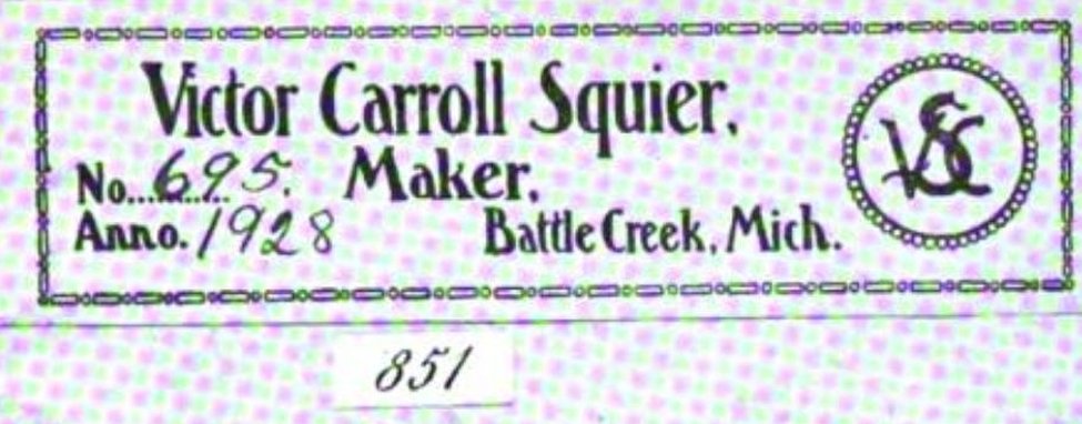 A printed violin label inside a plain rectangular frame of chain-link ornament. Bold condensed type reads Victor Carroll Squier on the first line, then Maker, then Battle Creek, Mich. Two blanks are completed by hand in ink: 695 after No., and 1928 after Anno. At the right, inside a beaded circle, a monogram interlaces the letters V, C and S. Below the frame the plate prints its facsimile number, 851, in a small ruled box. A coarse halftone dot screen from the 1930 printing lies over the whole image.